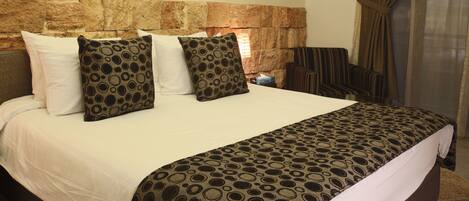Habitación de hotel con cabecera de piedra, una cama grande con ropa de cama estampada, dos cuadros colgados en la pared y vistas al exterior a través de una ventana con cortinas.