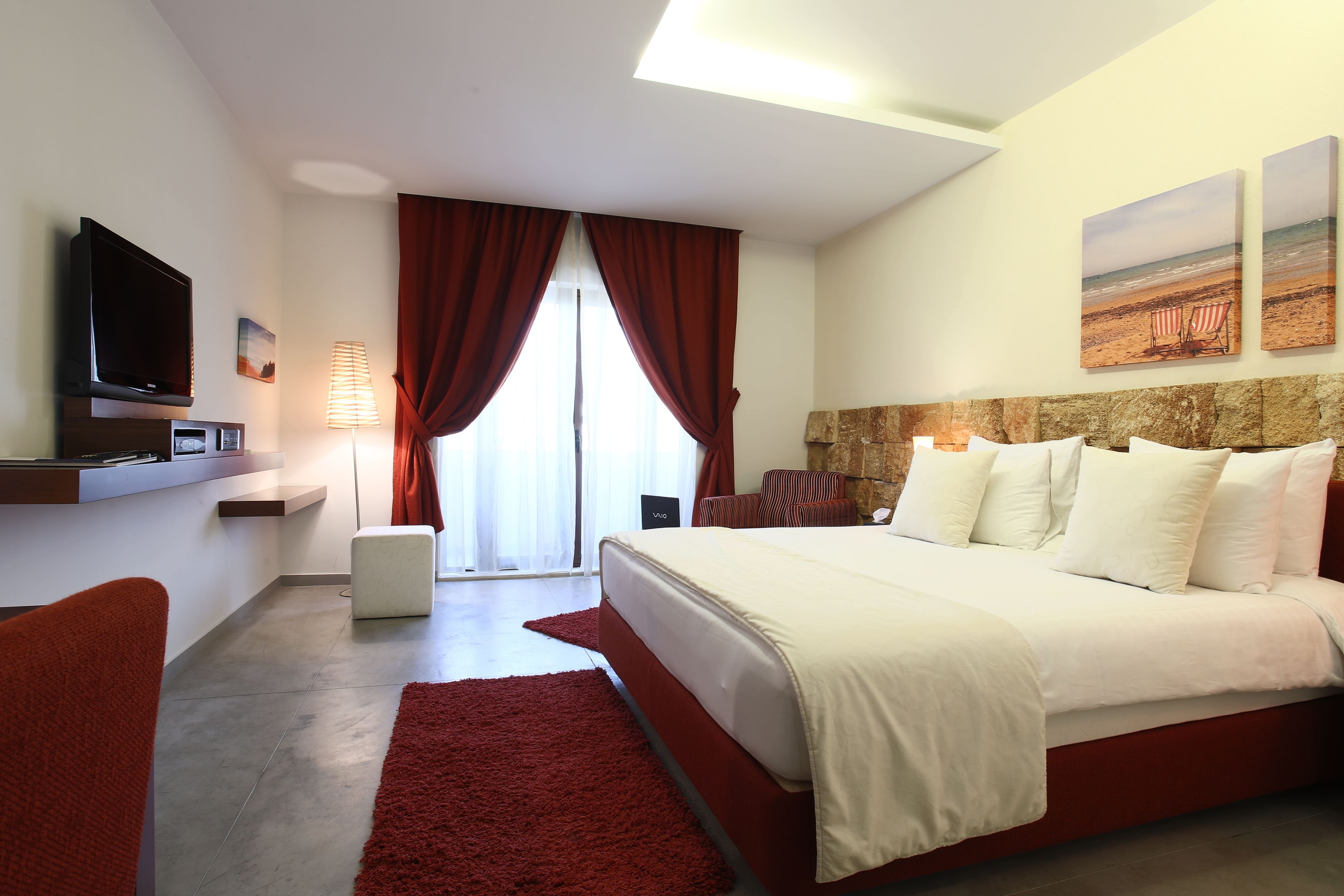 A modern hotel room with a large bed, a flat-screen TV, a desk with a lamp, and a view of the beach through the window.
