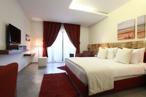 A modern hotel room with a large bed, a flat-screen TV, a desk with a lamp, and a view of the beach through the window. - Monoberge Byblos Hotel (Byblos)
