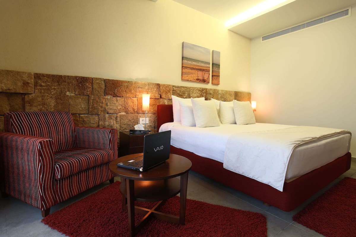 A hotel room with a bed, a chair, a desk with a laptop, a stone wall, and a painting on the wall.