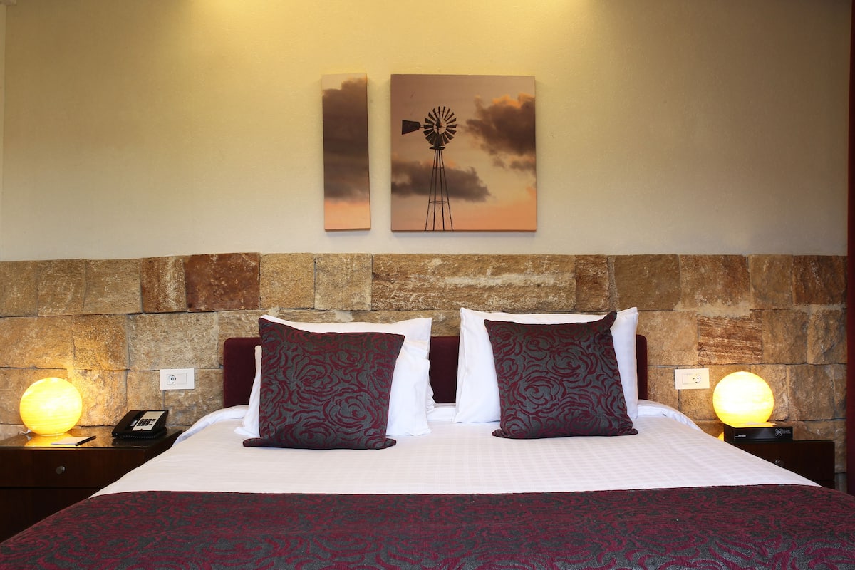 A neatly made bed with patterned pillows, a stone headboard, and two wall-mounted artworks depicting a windmill and a sunset.