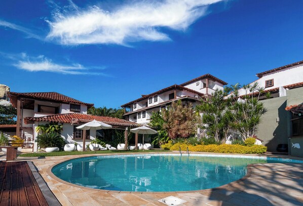 A pool area with a clear blue sky, a wooden deck, and a building with a tiled roof. - Arambaré Buzios Hotel (Búzios)