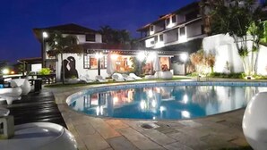 Vista nocturna de un hotel con piscina, sillones de descanso y un área exterior bien iluminada.
