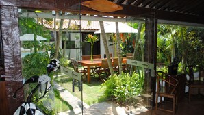 Une terrasse fermée par des baies vitrées, avec une table et des chaises en bois, entourée d’une végétation tropicale luxuriante.