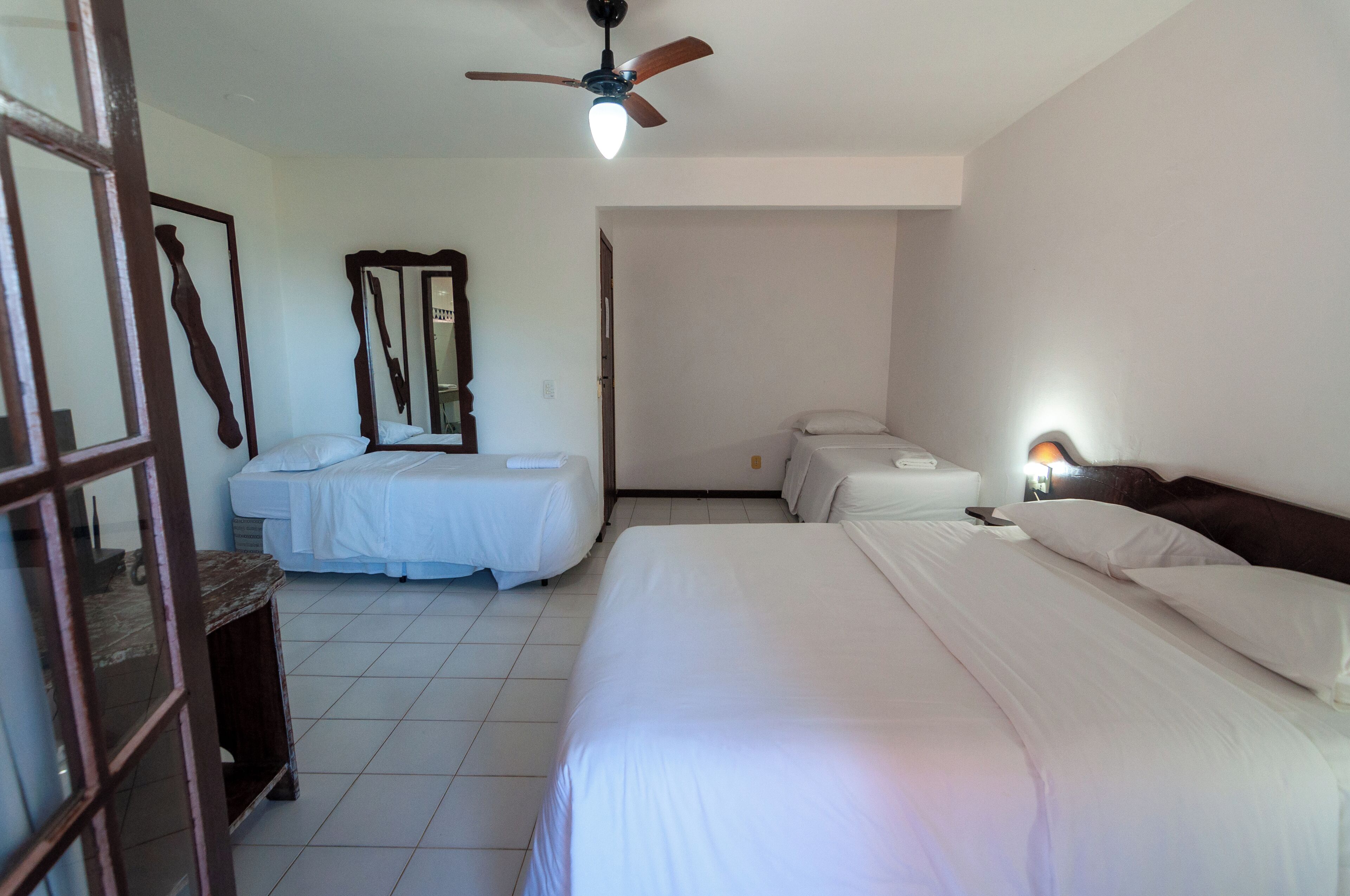 A hotel room with a large bed, a smaller bed, a ceiling fan, a bedside table with a lamp, and a mirror on the wall.