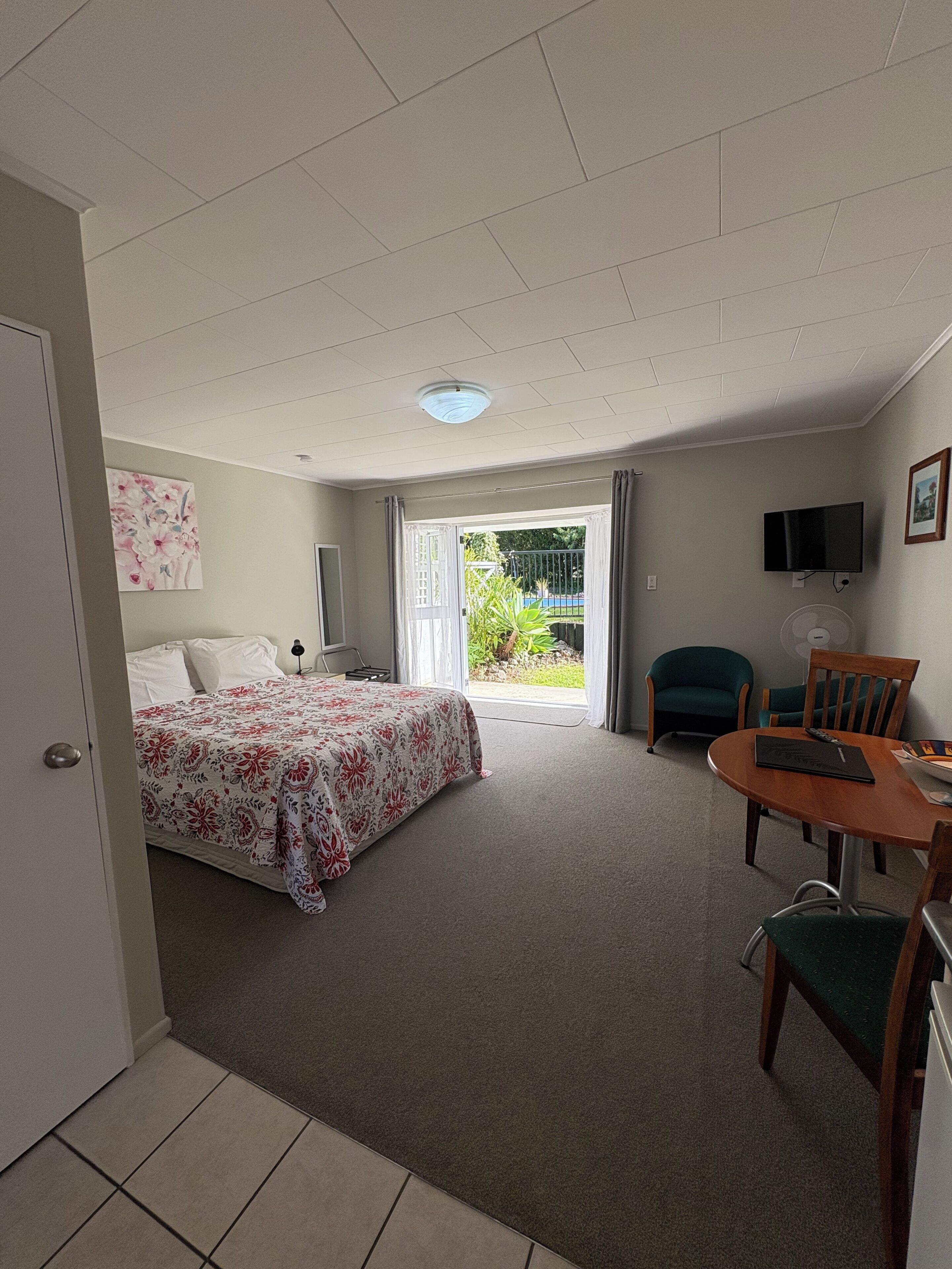 Une chambre à coucher avec un lit, un bureau et une chaise. On y trouve une porte qui donne sur une autre pièce, une fenêtre avec des rideaux et un luminaire au plafond.