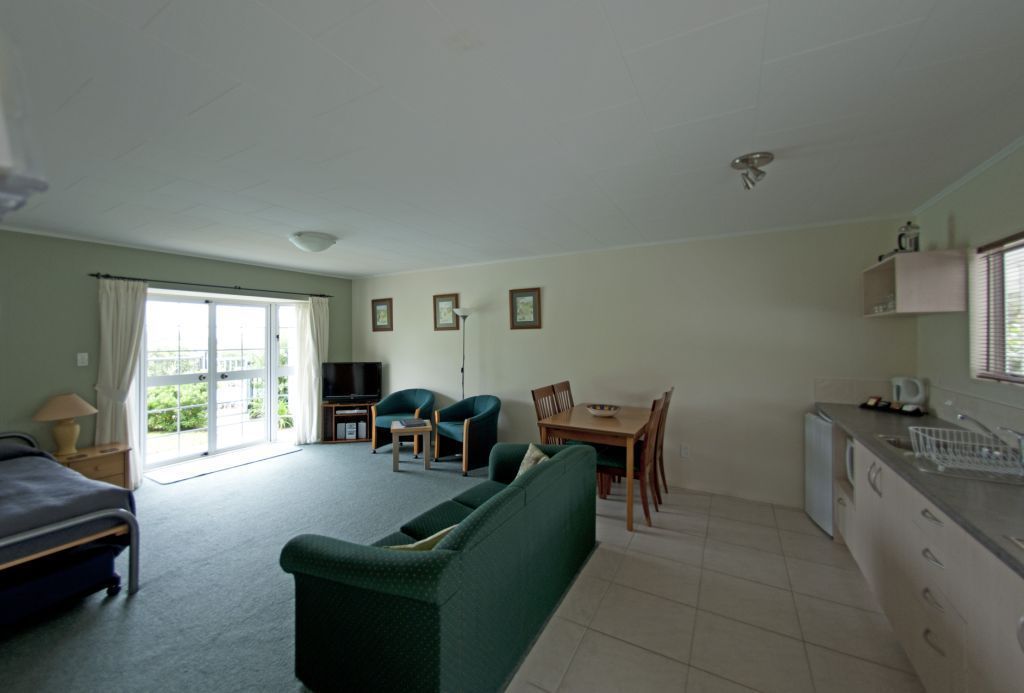 Een kamer met een bed, een groene bank, een eettafel met stoelen, een kitchenette en een schuifdeur van glas die leidt naar een balkon.