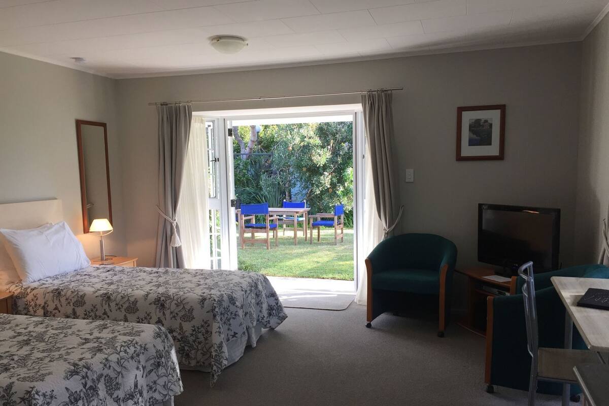 A hotel room with two beds, a desk with a chair, a television, and a view of a garden through an open door.