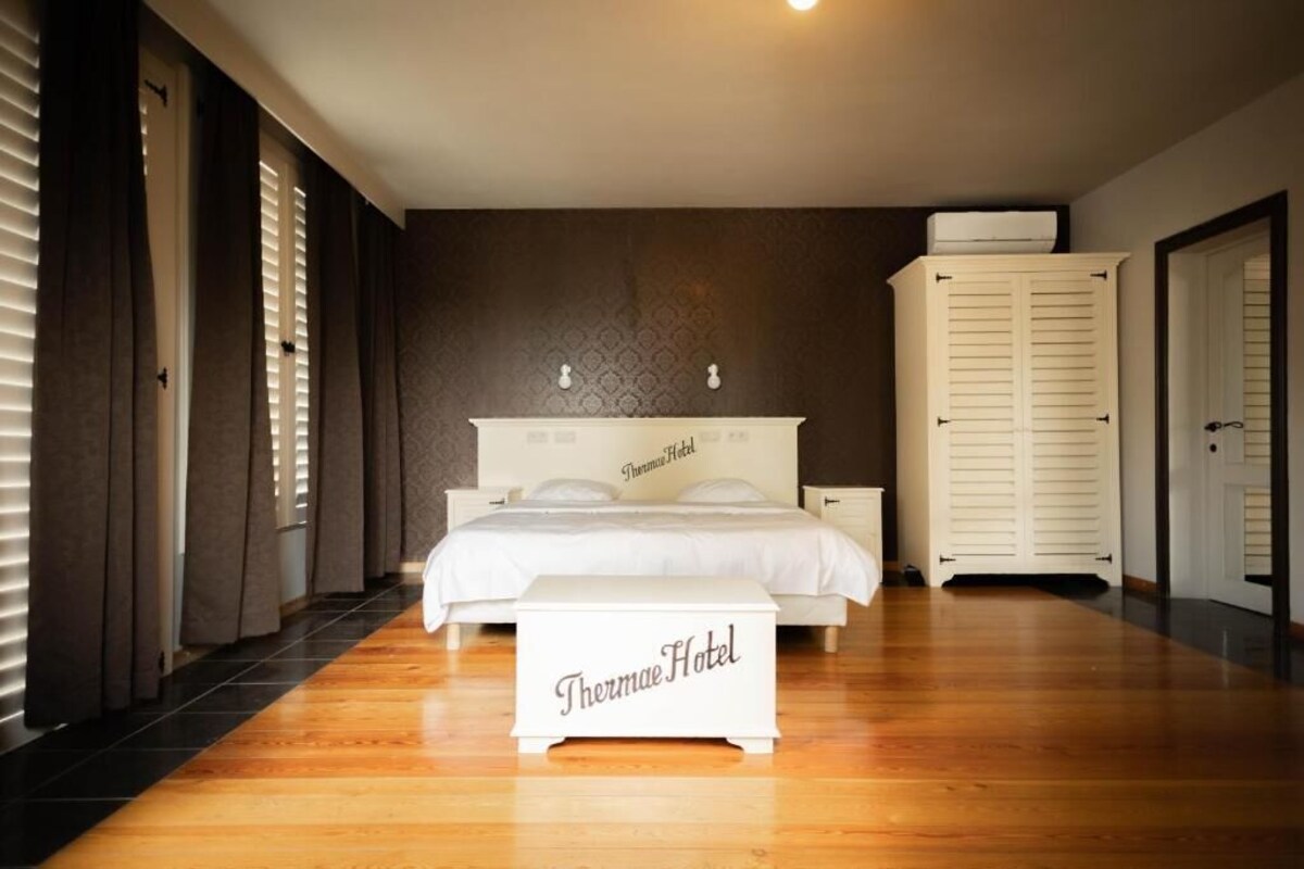 Een hotelkamer met een bed, nachtkastje, kledingkast en een bankje met de naam van het hotel.