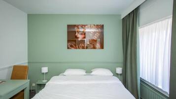 A hotel room with a bed, a desk, a chair, a wall-mounted artwork, and a window with curtains.