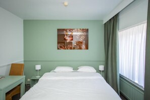 Habitación de hotel con cama, escritorio, silla, una obra de arte en la pared y ventana con cortinas.