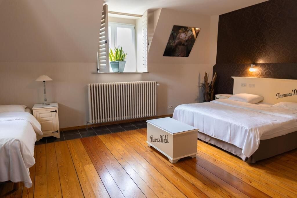 A hotel room with a bed, bedside table, nightstand, radiator, window with curtains, and a small plant.