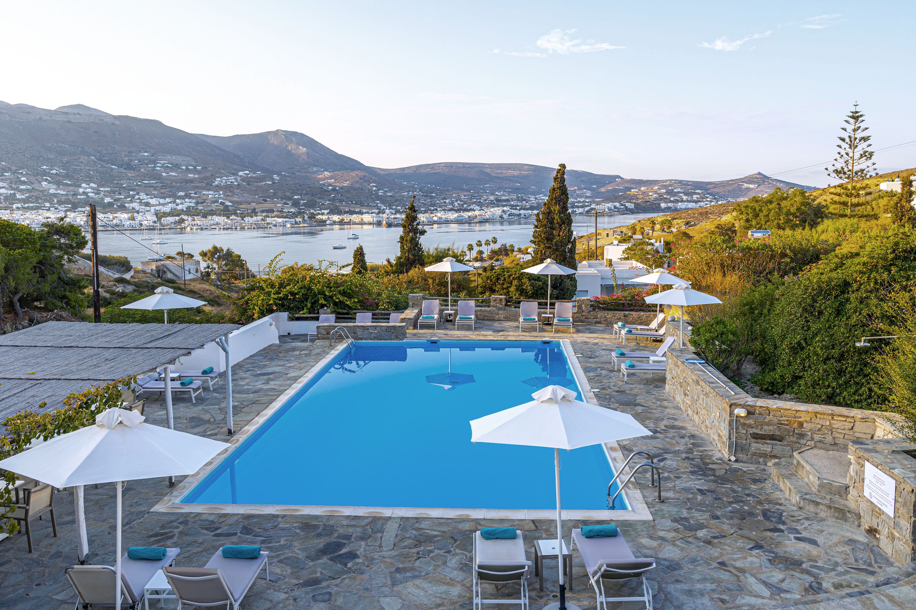 Piscina rectangular con solárium de piedra, rodeada de sillas de descanso y sombrillas, con vistas a un pueblo costero y colinas.