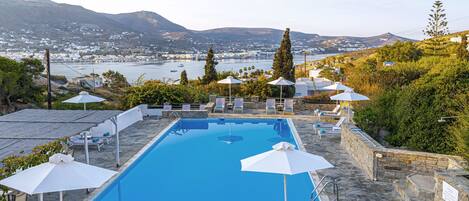 Piscina rectangular con solárium de piedra, rodeada de sillas de descanso y sombrillas, con vistas a un pueblo costero y colinas.