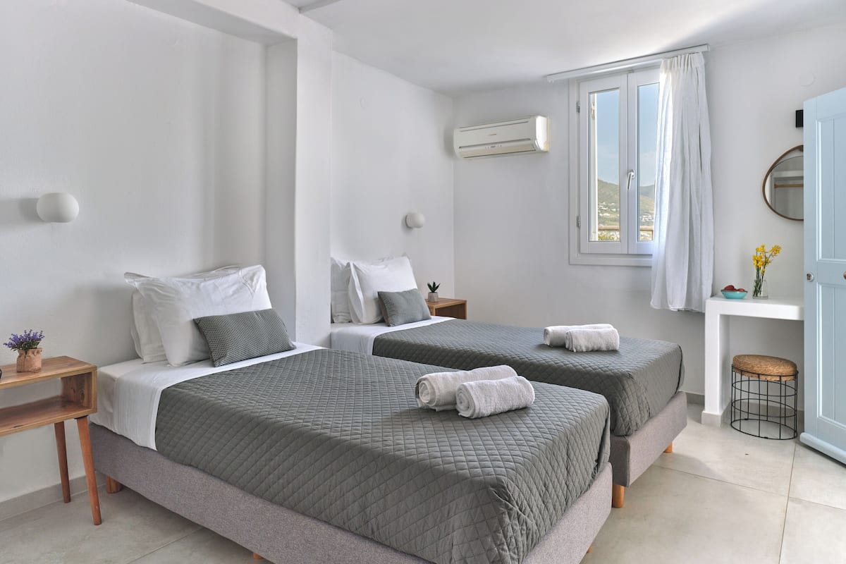A hotel room with two beds, a small desk, a window with curtains, and a wall-mounted air conditioner.