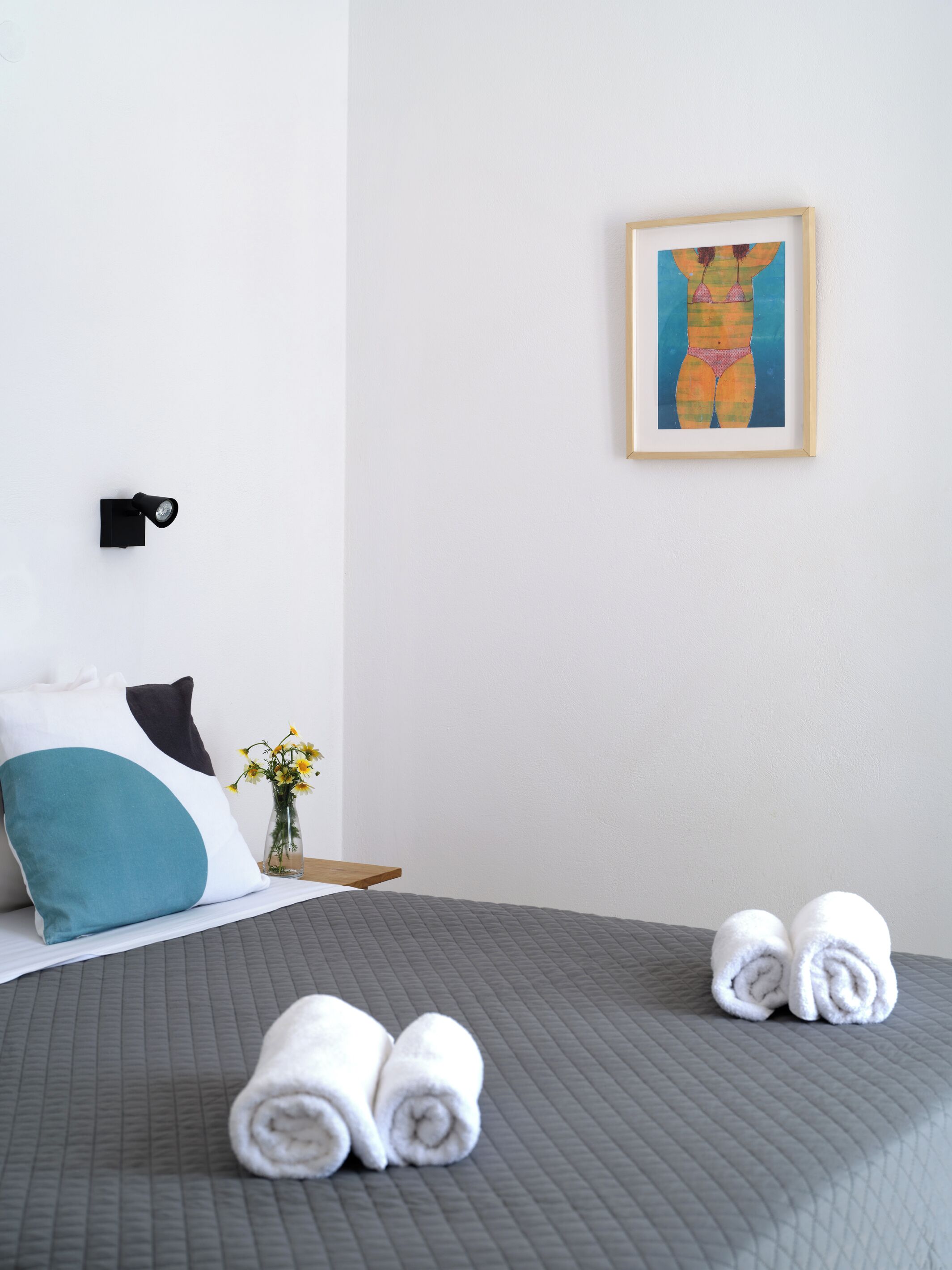 A neatly made bed with grey bedding, two rolled towels, a teal and white pillow, a black wall-mounted light, and a framed artwork on the wall.