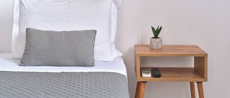 A neatly made bed with a patterned pillow and a plain pillow, a bedside table with a remote control and a small potted plant, and a wall-mounted light fixture.