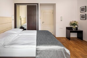 A hotel room with a bed, a bedside table with a vase, and a door leading to another room.