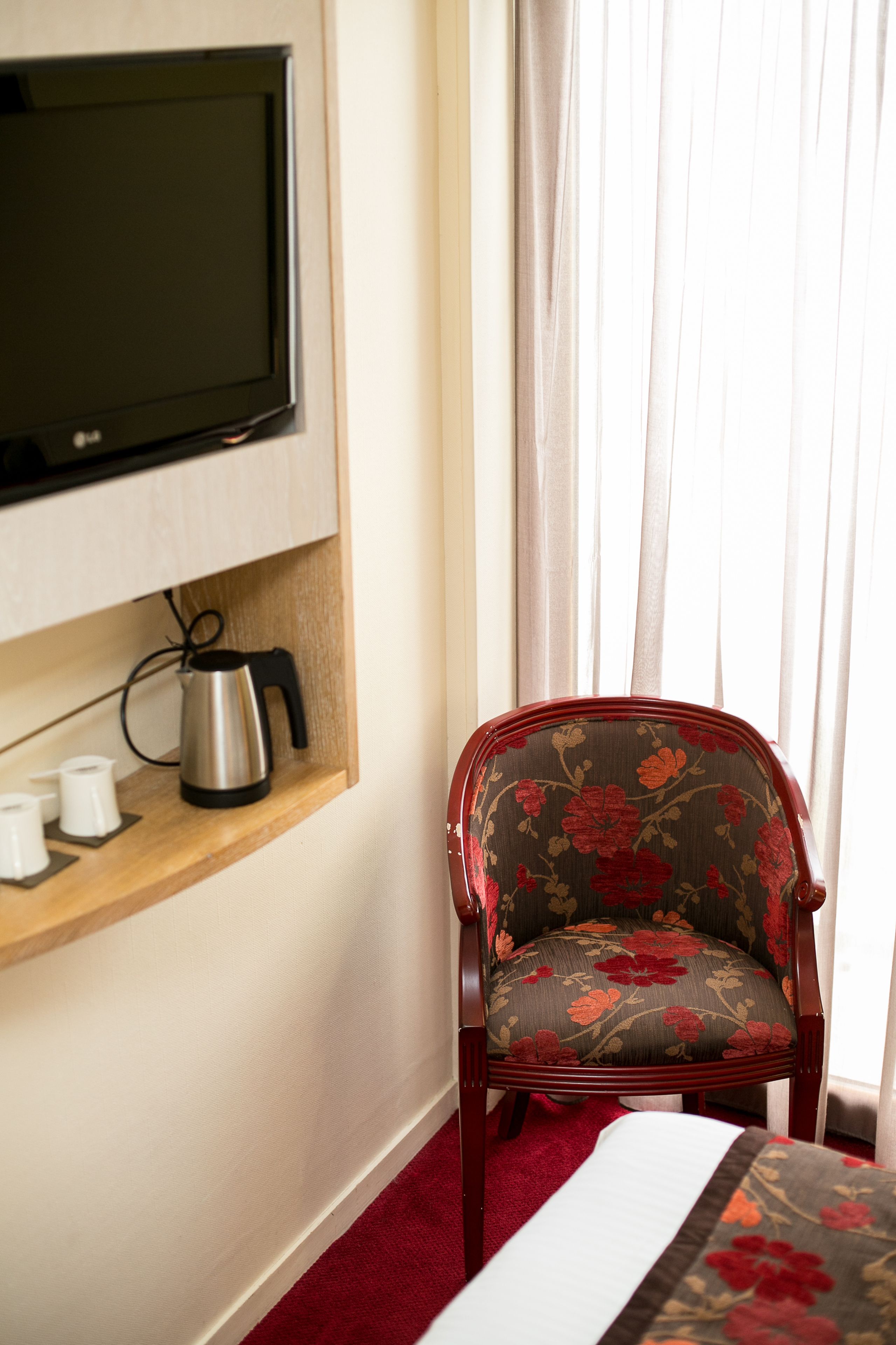 Hotelkamer met een aan de muur bevestigde televisie, een houten plank met waterkoker, bekers en een bloemenstoel.