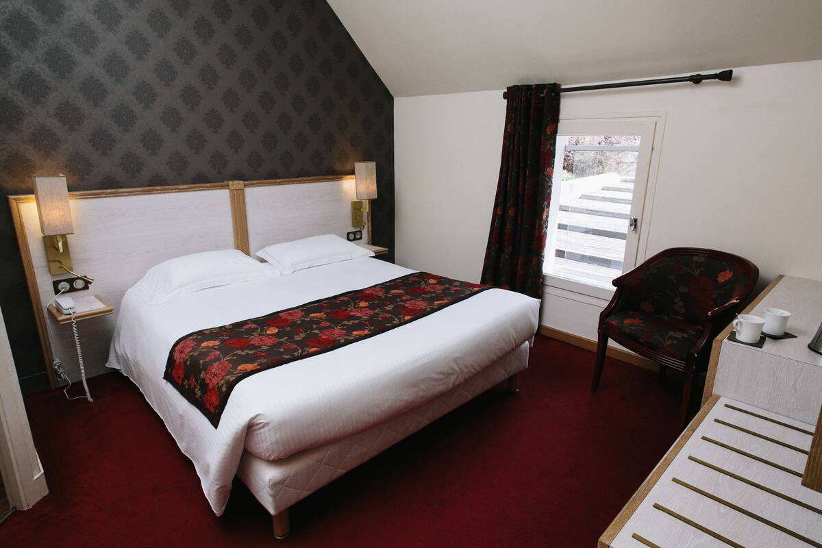 Ein Hotelzimmer mit einem großen Bett, einem Schreibtisch mit Stuhl, einem Telefon und einem Fenster mit Vorhängen.