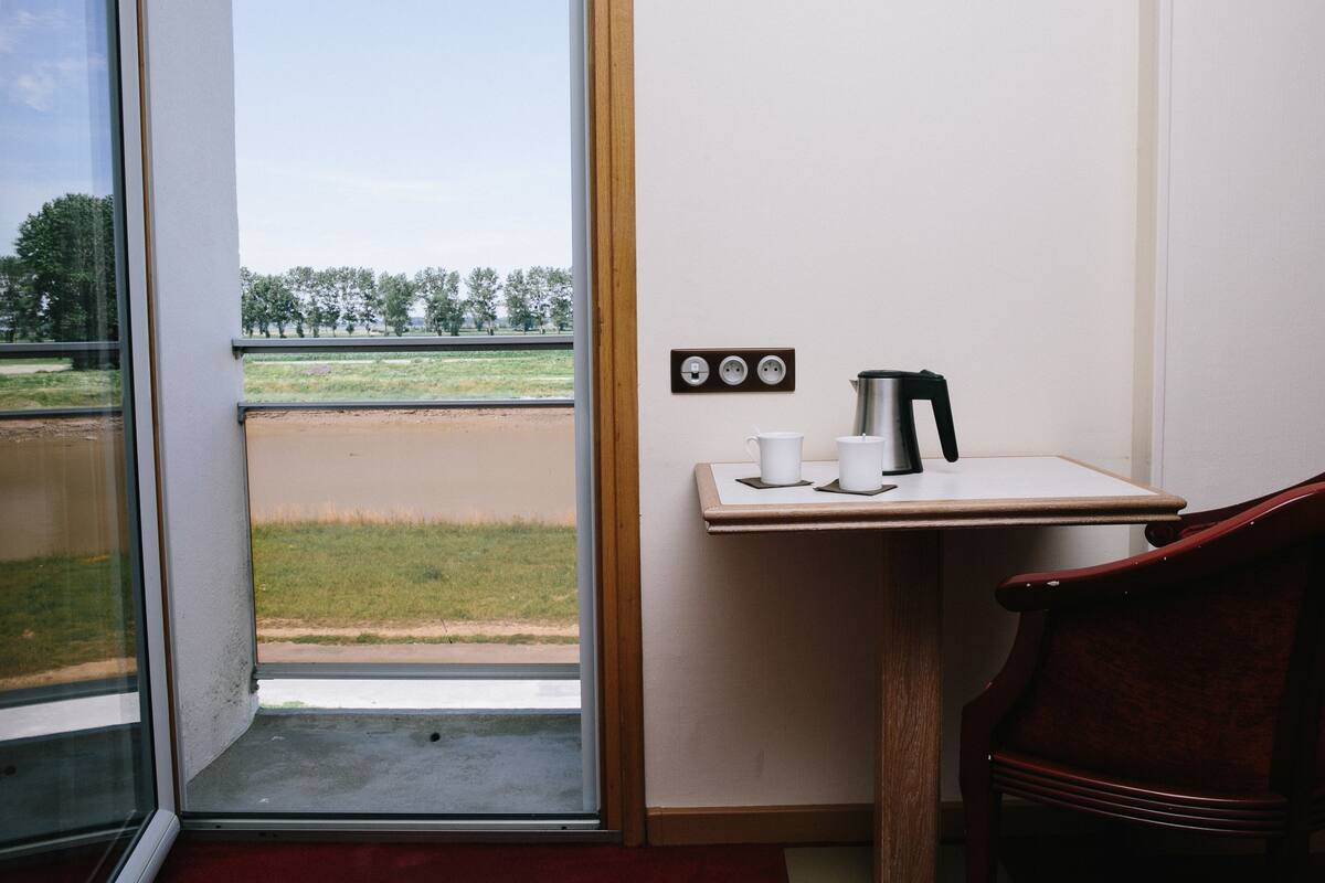 Ein Zimmer mit Fenster, einem kleinen Tisch mit Wasserkocher und Tassen sowie einem Stuhl.
