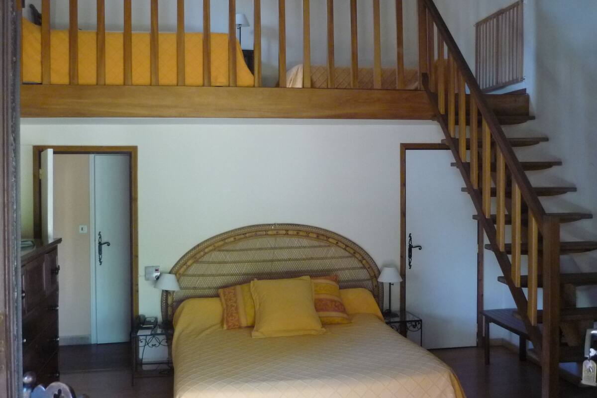A bedroom with a bunk bed, a wooden staircase, a bedside table with a lamp, and a door leading to another room.