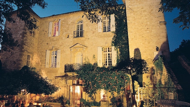 A historic stone building with multiple windows and balconies, surrounded by trees and illuminated by warm lighting.