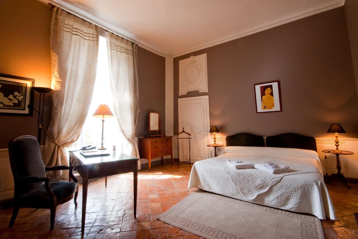 A hotel room with a bed, a desk, a chair, a dresser, and a window with curtains.