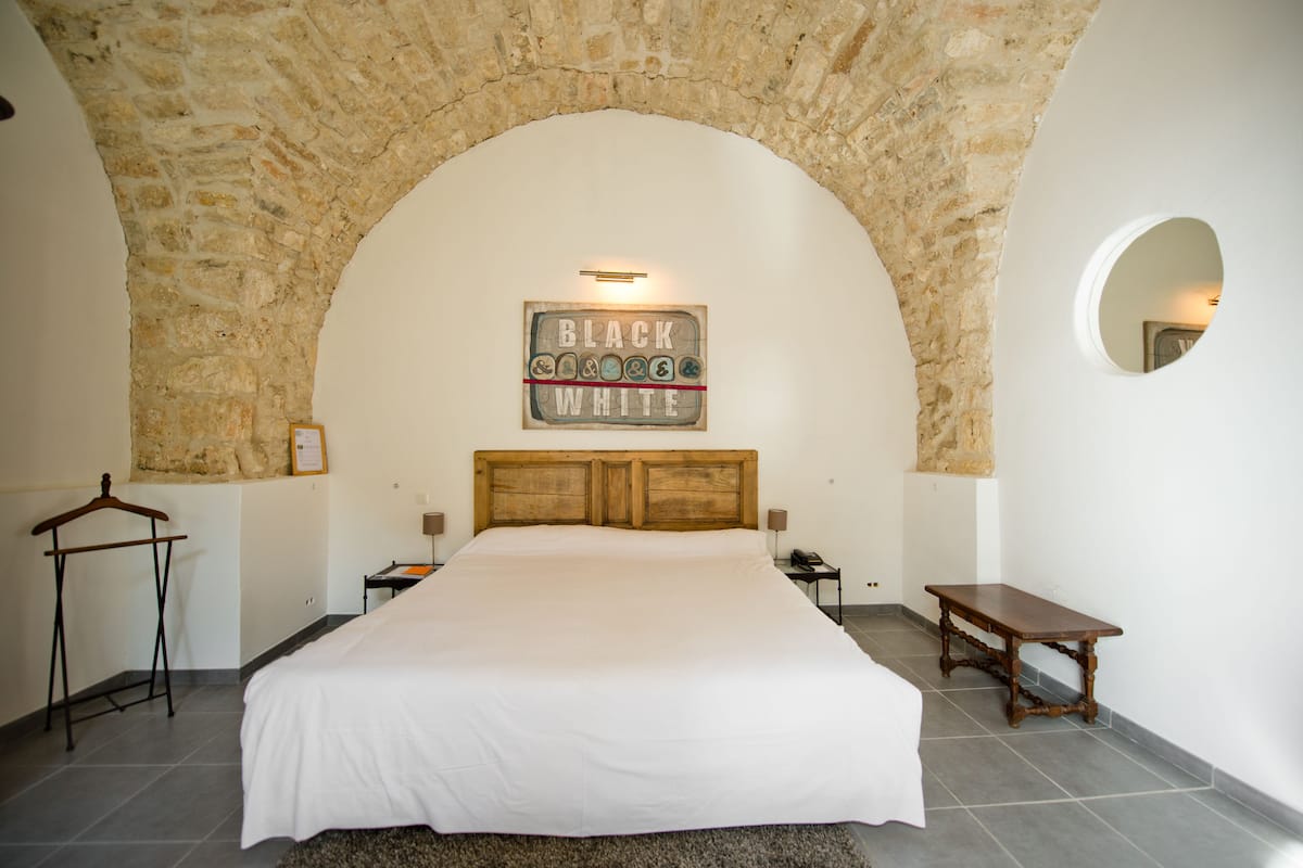 A bedroom with a stone arch ceiling, a large bed, a wooden headboard, a bedside table, a nightstand, a round mirror, and a sign above the bed.
