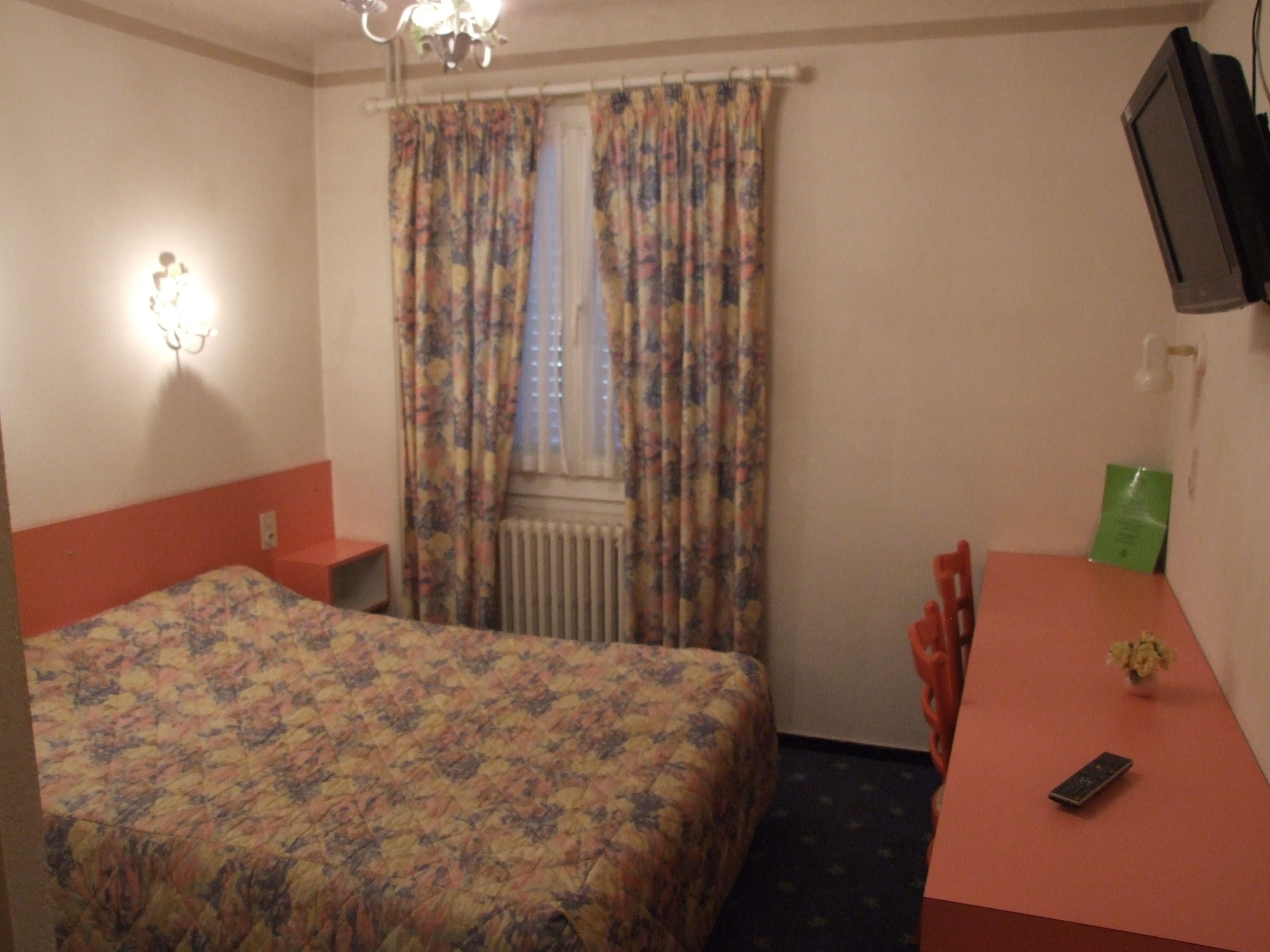 A hotel room with a bed, a desk, a chair, a TV, and a radiator.