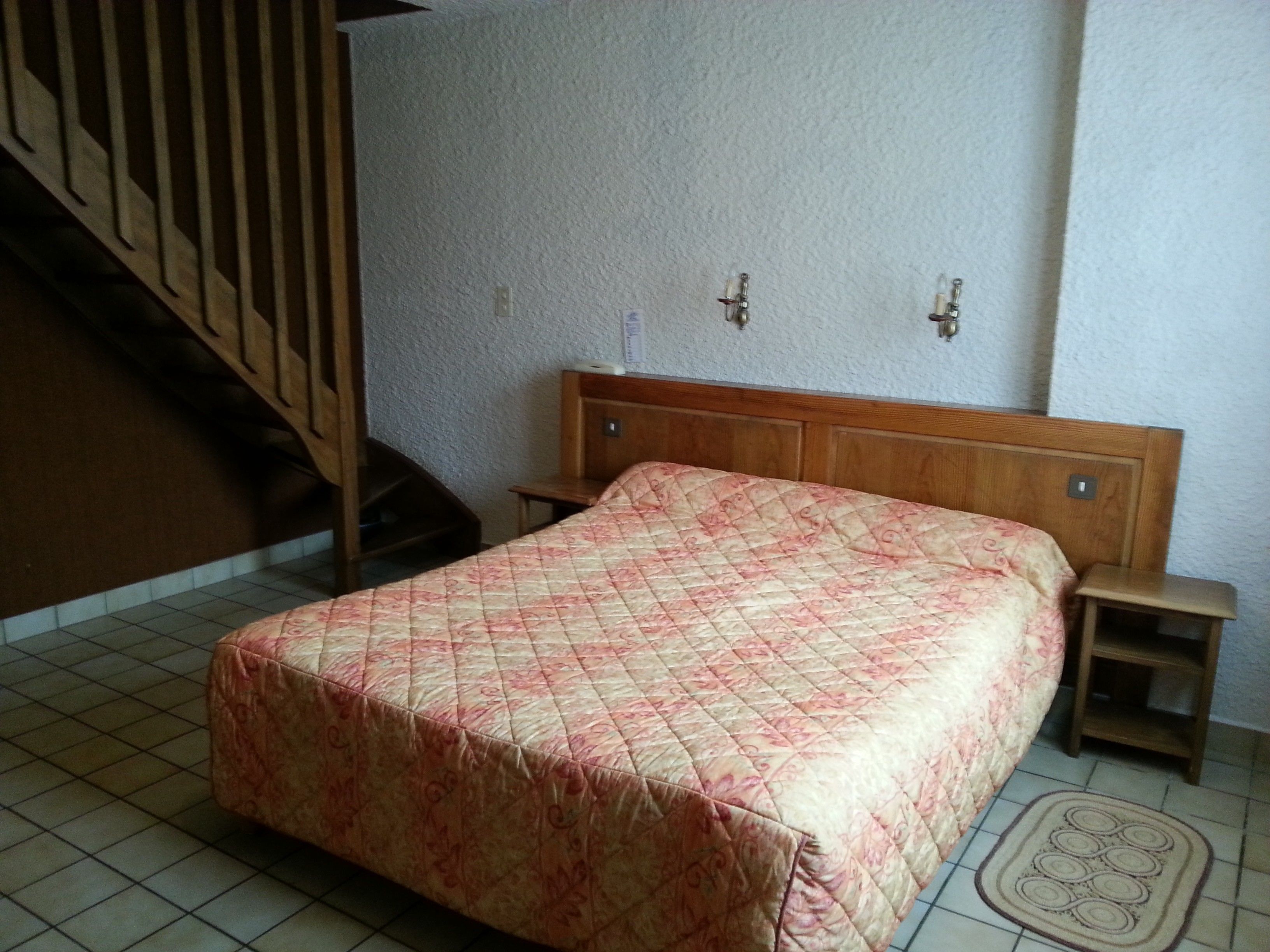 A bedroom with a bed, a nightstand, a wooden headboard, a wall-mounted light fixture, and a staircase.