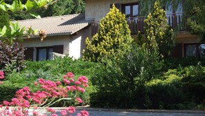 A two-story building with a tiled roof, surrounded by lush greenery and flowering plants.