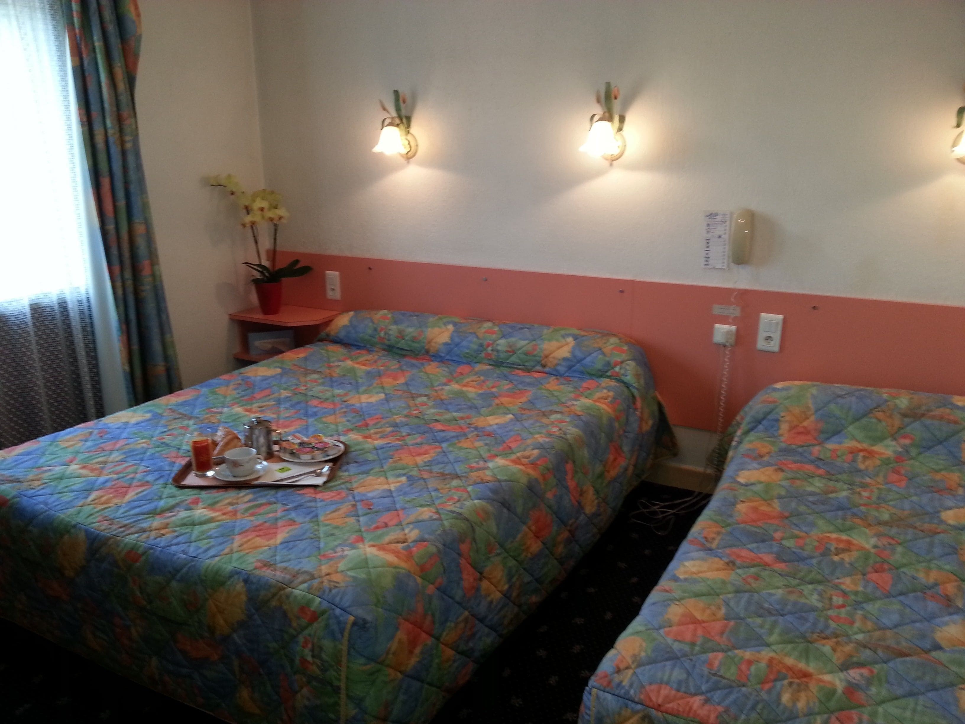 A hotel room with two beds, a small table with breakfast items, a potted plant, and wall-mounted lights.