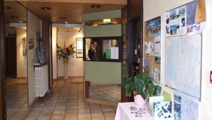 A reception area with a reception desk, a bulletin board with various notices, a potted plant, and a tiled floor.