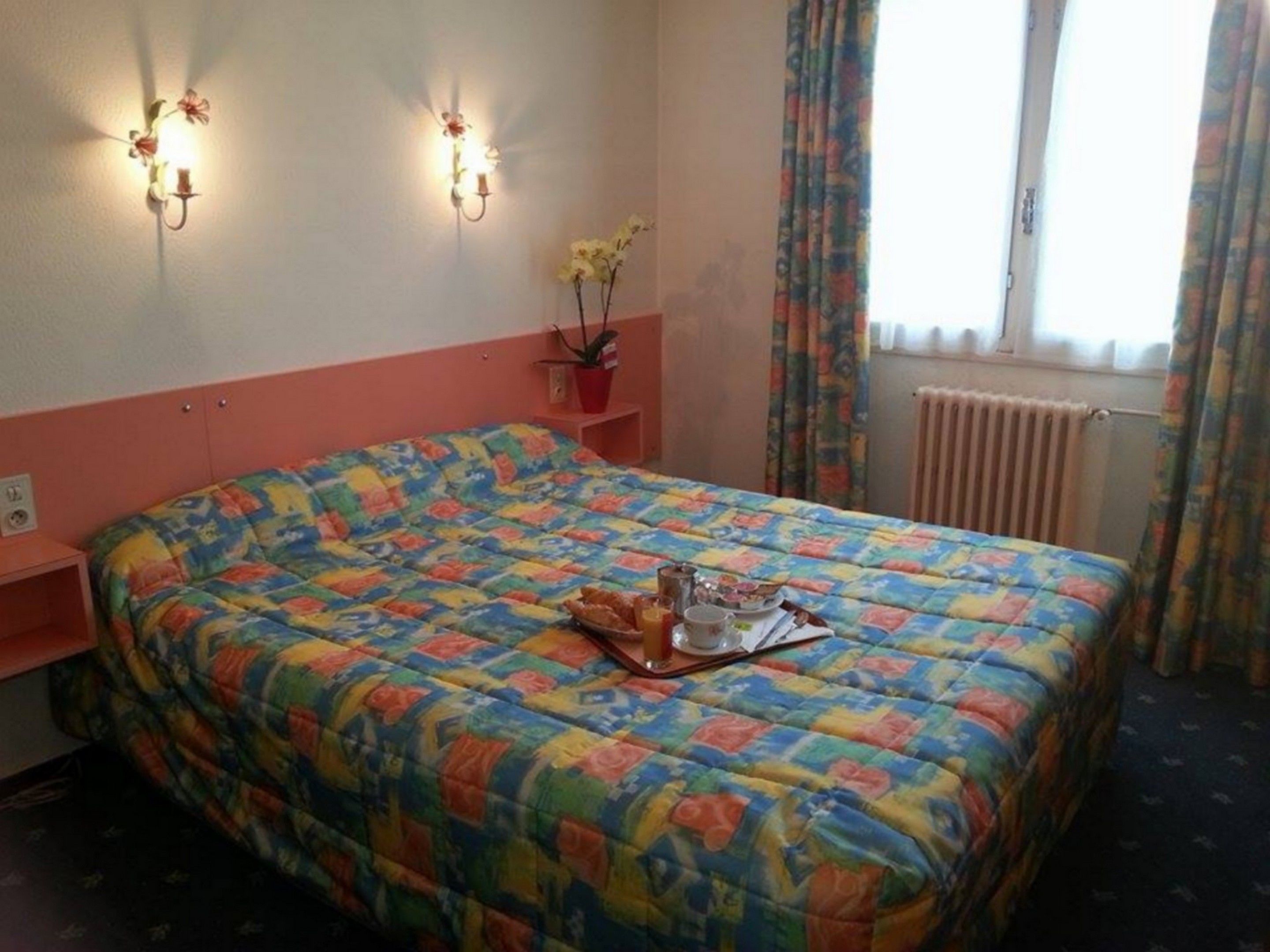 A bedroom with a colorful bedspread, a bedside table with a lamp, a potted plant, and a window with curtains.