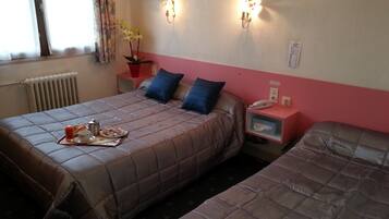 A hotel room with two beds, a bedside table with a lamp, a radiator, and a window with curtains.
