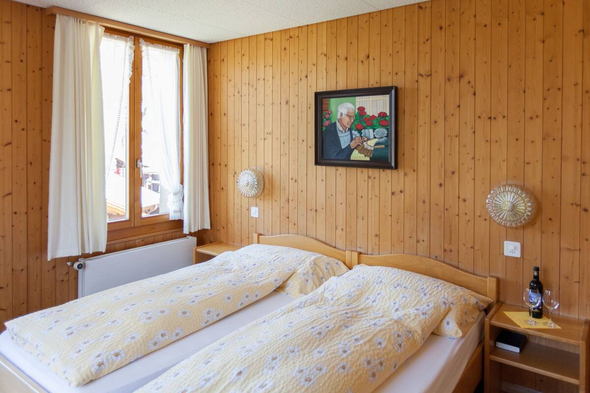 Una habitación con paredes de paneles de madera, dos camas individuales con ropa de cama floral, un cuadro en la pared y una mesita de noche con una botella y vasos.