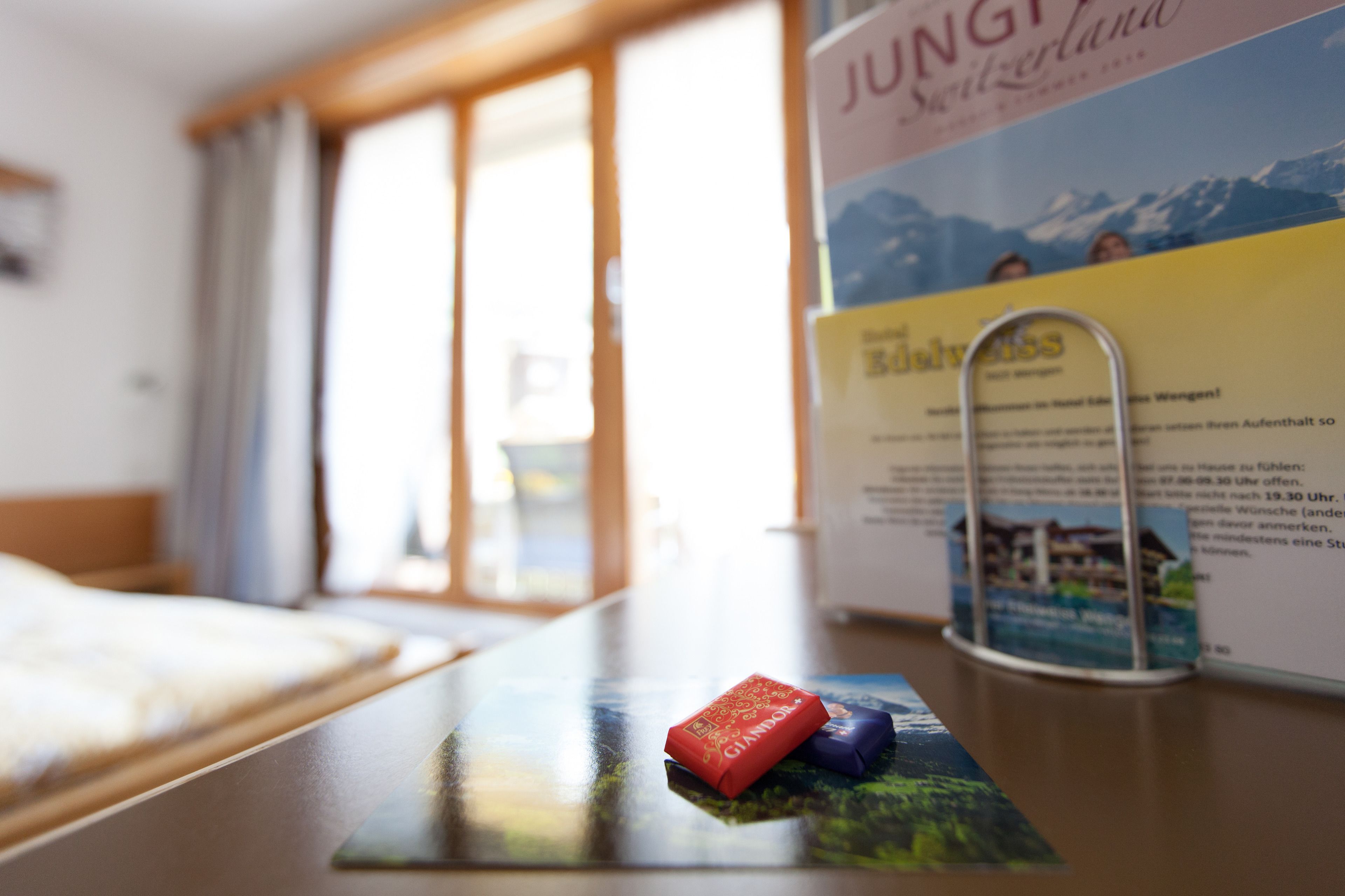 A hotel room with a bed, a window with curtains, and a table with a brochure and a pen holder.