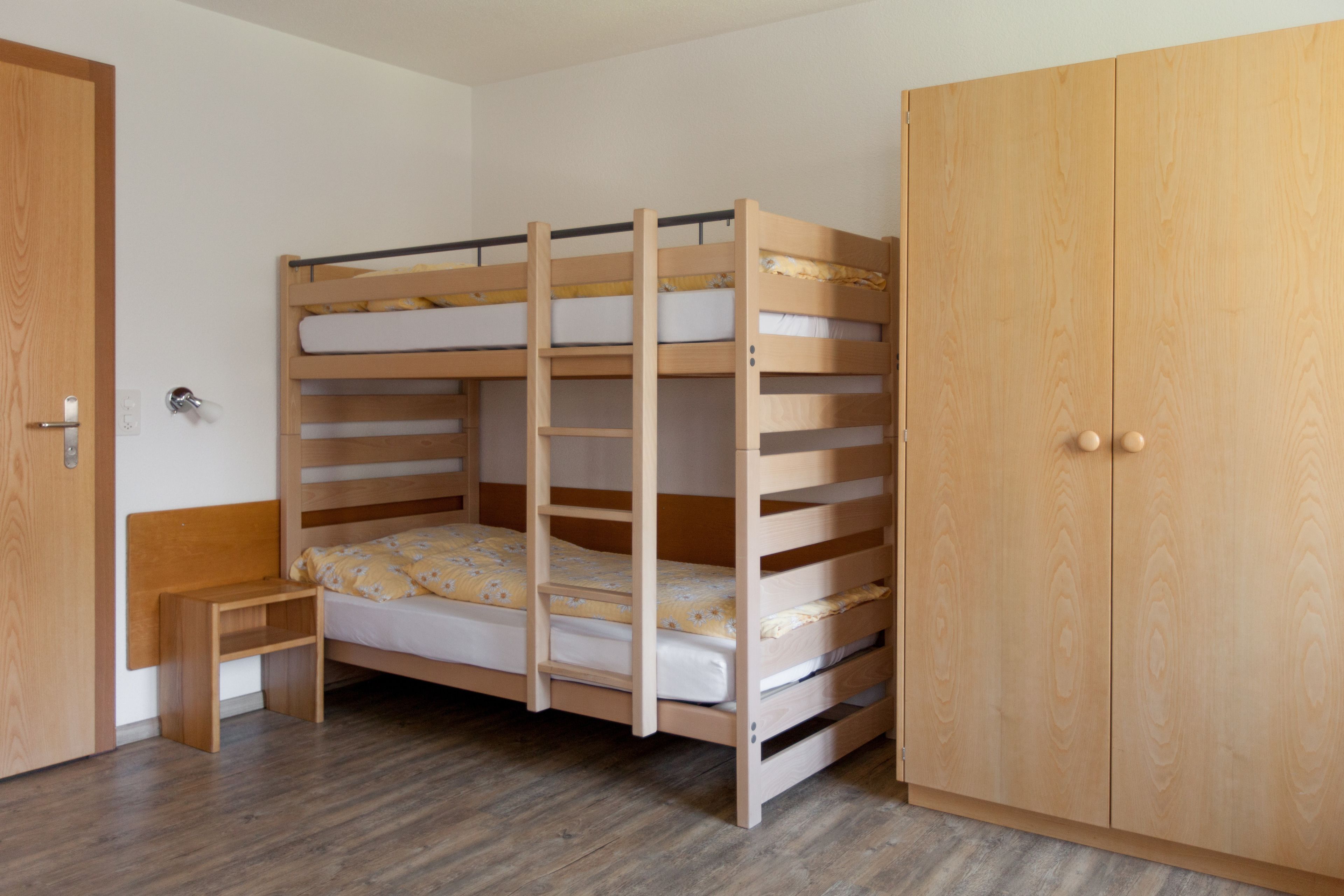 Ein Stockbett mit Holzbettrahmen und eine kleine Bank daneben. Ein zweitüriger Holzschrank steht ebenfalls dort.