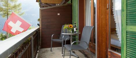 Un balcon orné d’un drapeau suisse, une table avec des chaises et un vase de fleurs.
