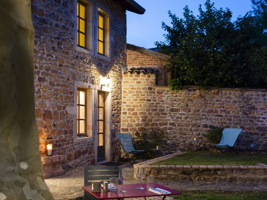 Ein Steinhaus mit Tür und Fenstern, ein Tisch mit Gläsern und einer Kerze sowie zwei Stühle davor.