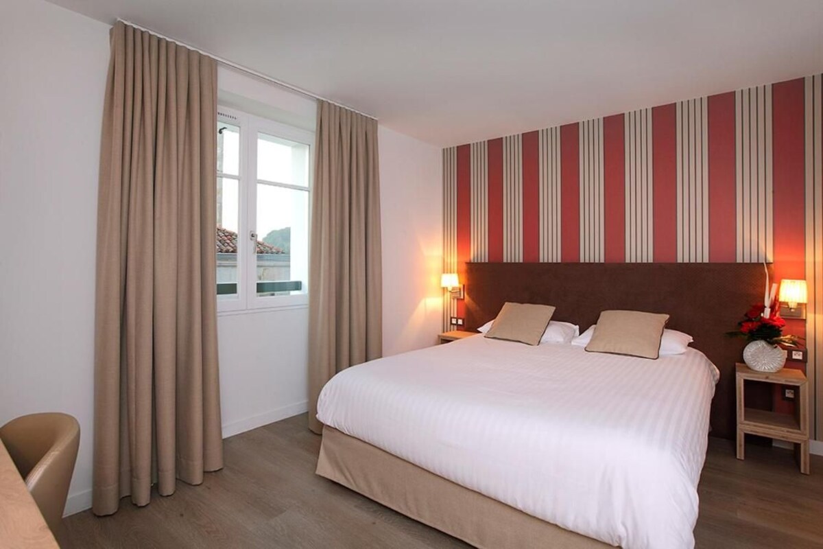 Una habitación de hotel con una cama grande, dos mesitas de noche con lámparas, un ventanal con cortinas y una pared a rayas.