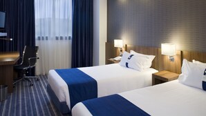 Ein Hotelzimmer mit zwei Betten, einem Schreibtisch und einem Sessel. Zwei Nachttische mit Lampen sind vorhanden.
