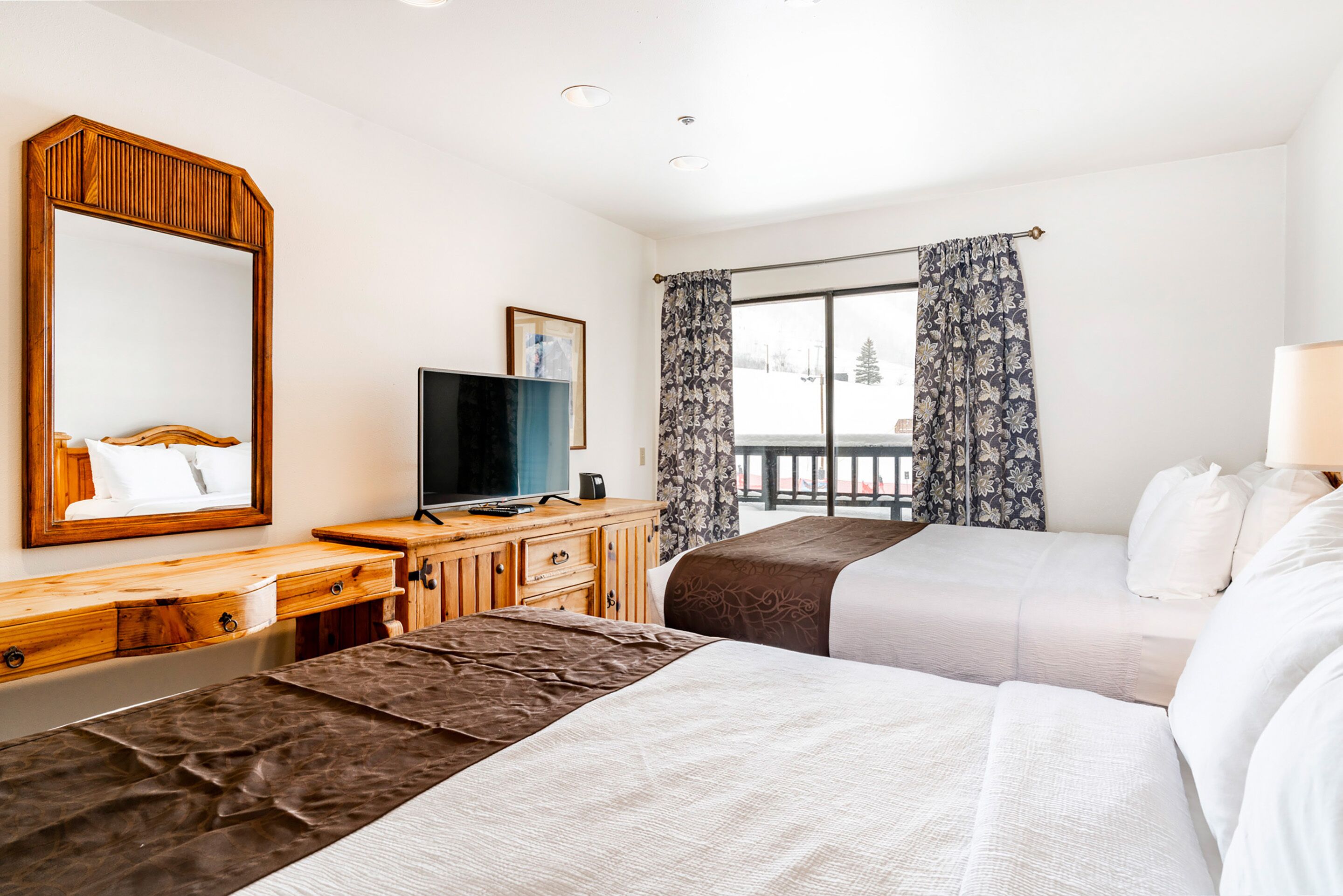 A hotel room with two beds, a wooden dresser, a television, and a large window with curtains.