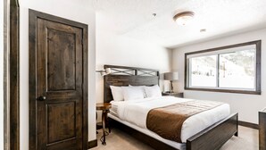 A bedroom with a large bed, a wooden headboard, a nightstand, a lamp, and a window with a view of a snowy landscape. - The Loft at Mountain Village by All Seasons Resort Lodging (Park City)