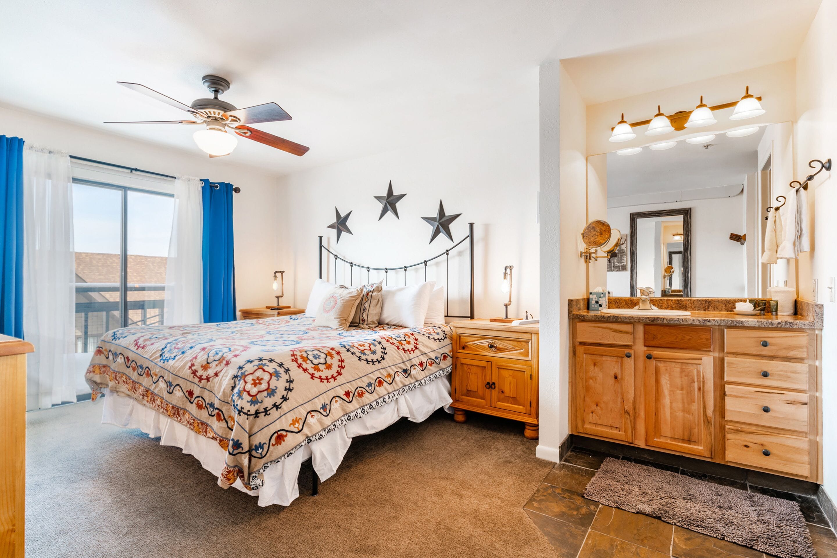 A bedroom with a bed, a nightstand, a dresser, a ceiling fan, and a bathroom with a vanity and mirror.