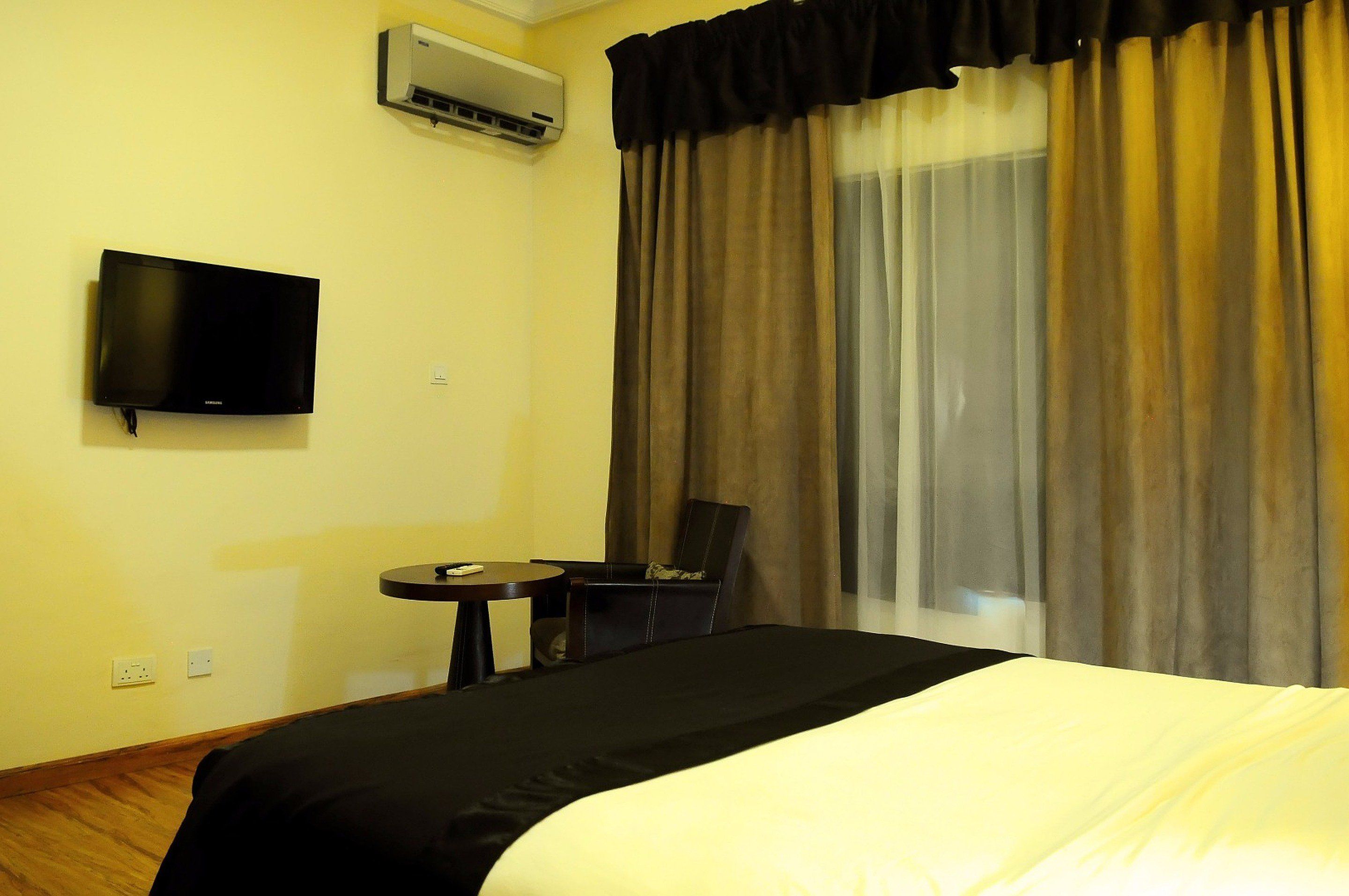 A hotel room with a bed, a television mounted on the wall, a small table, a chair, and a window with curtains.