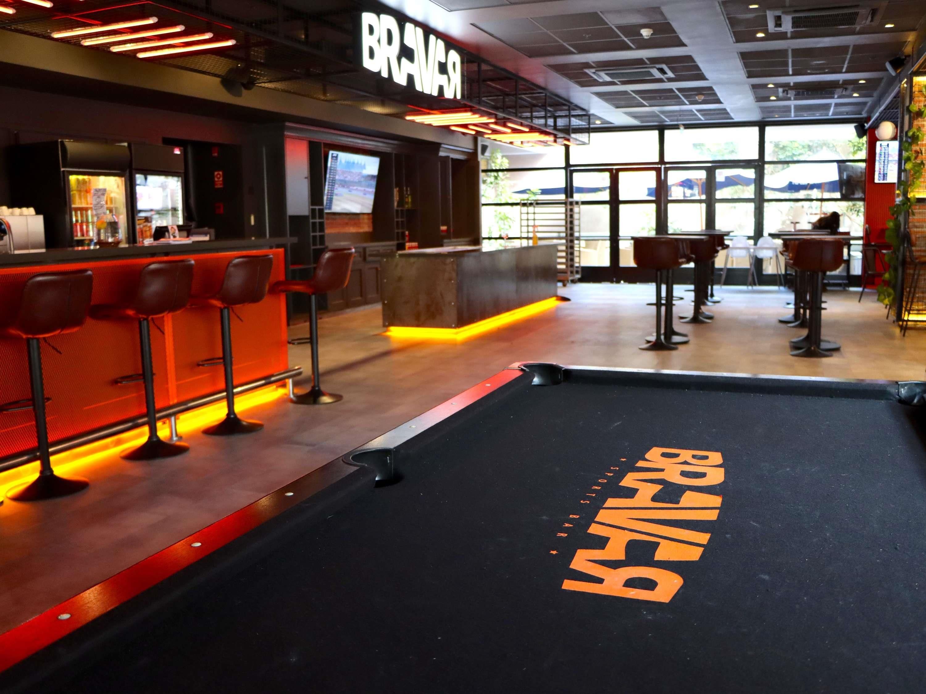 Una mesa de billar con la inscripción 'BRAVO', una zona de bar con taburetes y un ventanal grande al fondo.