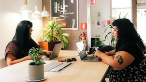 A modern office space with two individuals seated at a table with laptops, surrounded by potted plants and office supplies.