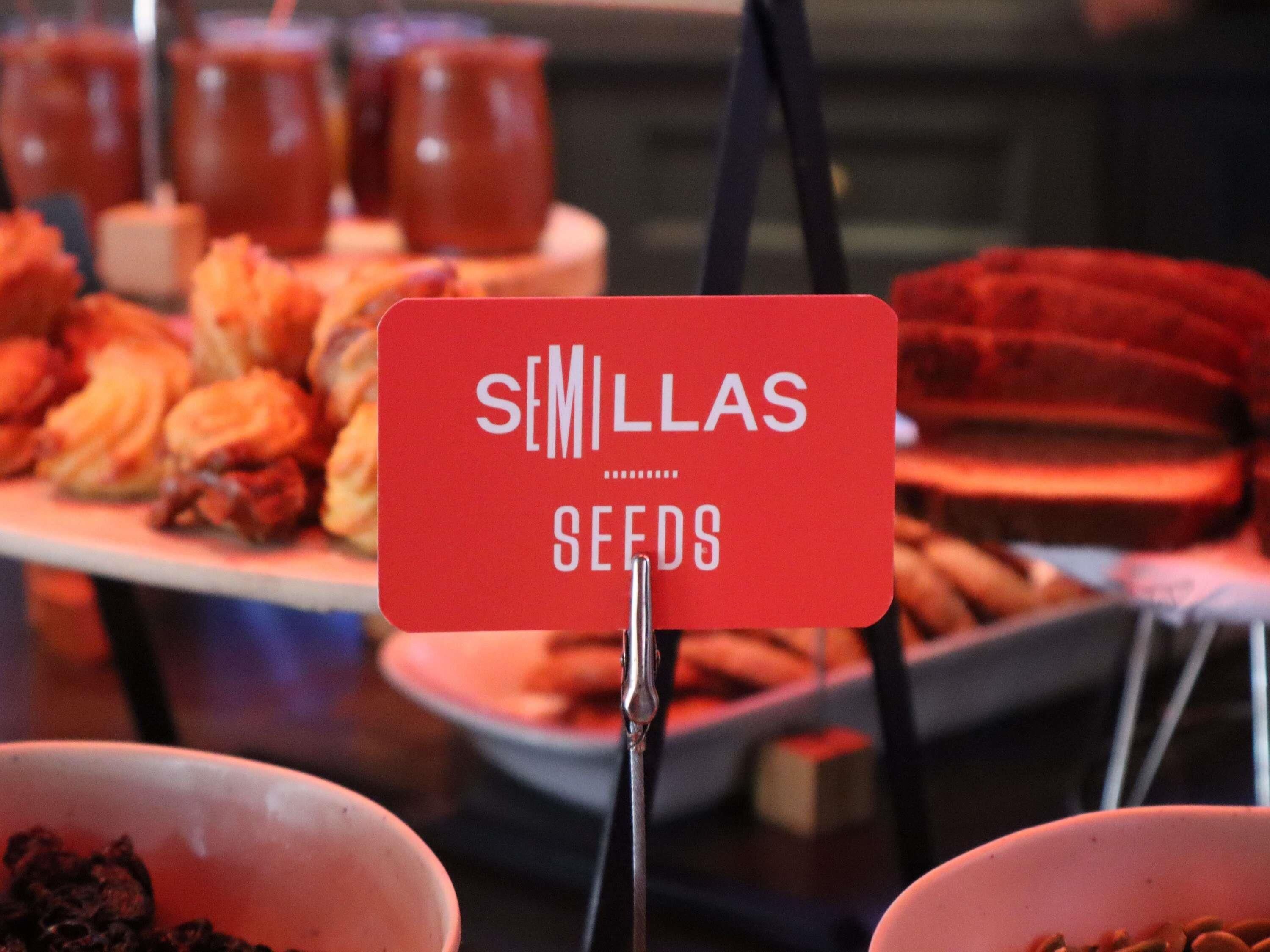 A sign with the text 'SEMINAS SEEDS' in a red square with a white border, placed in front of a display of various food items.